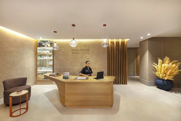 A reception desk with a receptionist, a wooden counter, a seating area with a chair and stool, and a decorative plant. - Pestana Cr7 Marrakech (Marrakech)