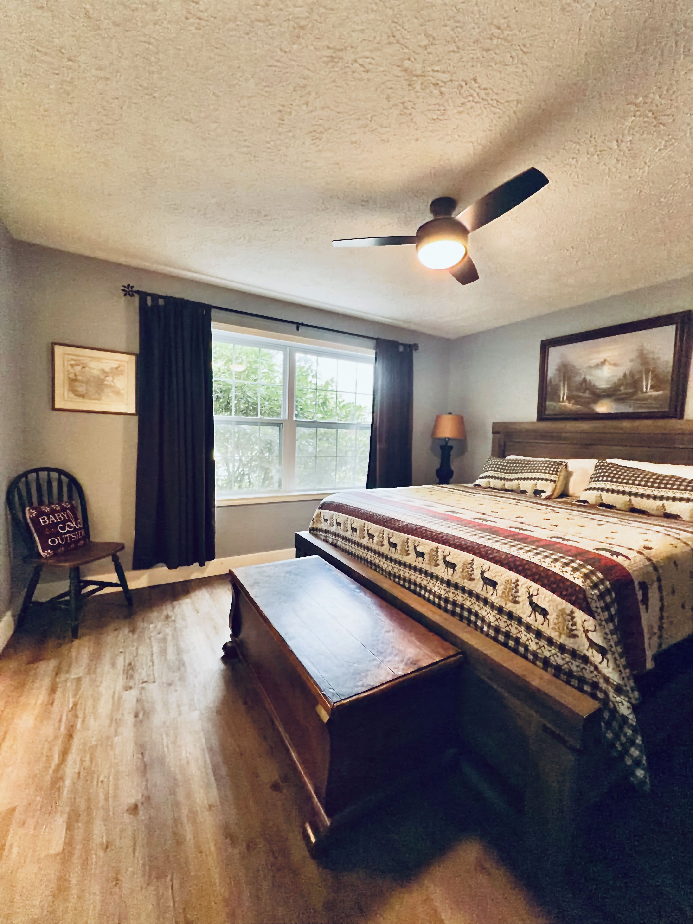 A bedroom with a bed, a chair, a nightstand, a ceiling fan, and a window with curtains.