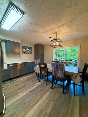 A modern kitchen with built-in appliances, a dining area with wooden chairs and a table, and a large window with a view of greenery.