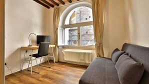 A room with a wooden floor, a desk with a chair, a grey sofa, and a large arched window with curtains. - Independent apartment near Piazza del Campo (Siena)