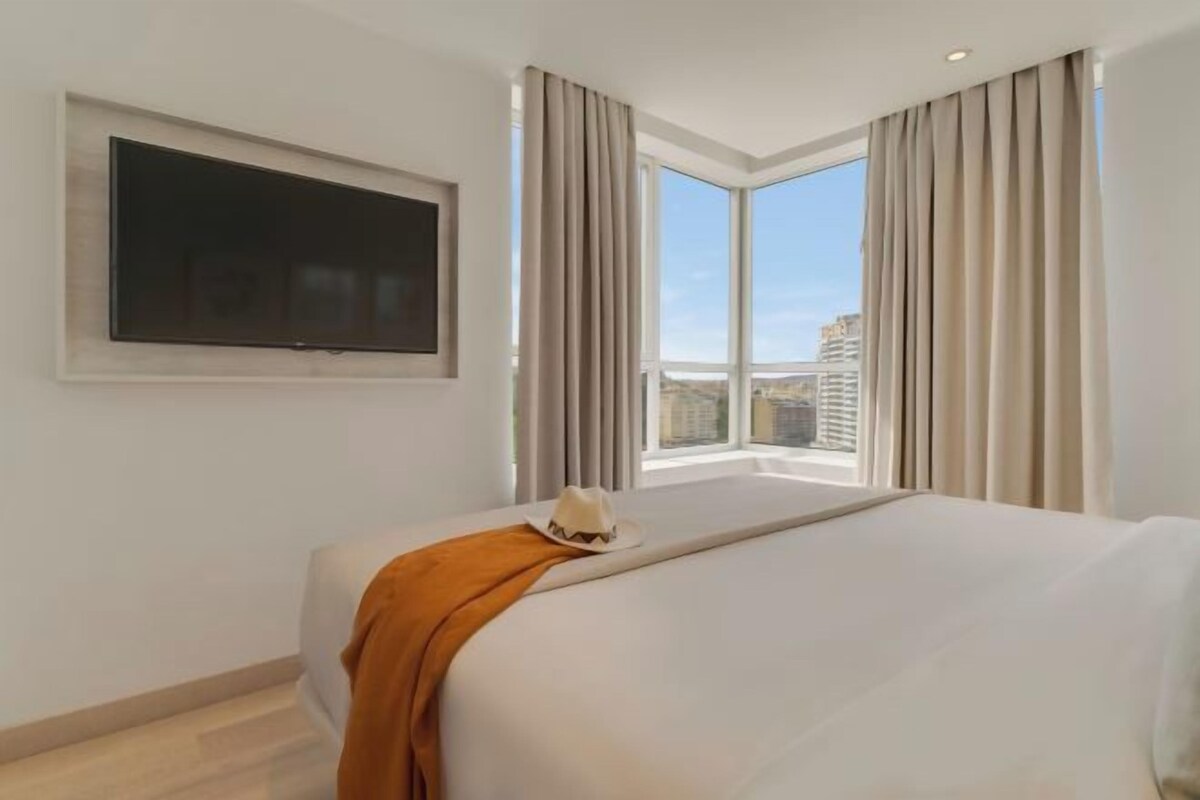 Una habitación de hotel con una cama grande, un televisor de pantalla plana montado en la pared y una ventana con cortinas.