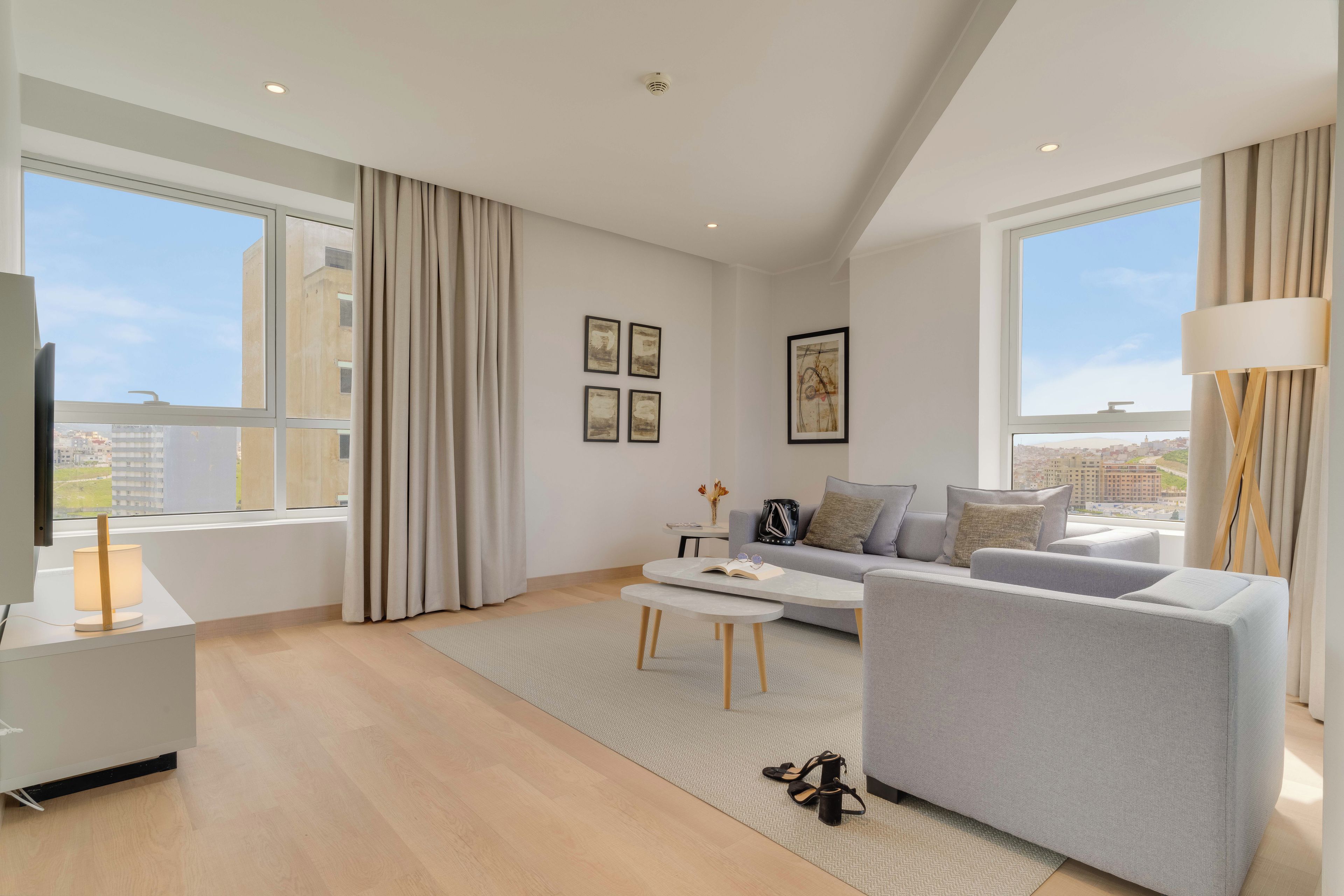 Una sala de estar moderna con un sofá gris, una mesa de centro y un televisor sobre un mueble bajo. Hay ventanas amplias con cortinas, vistas a la ciudad y una lámpara sobre una mesita auxiliar.
