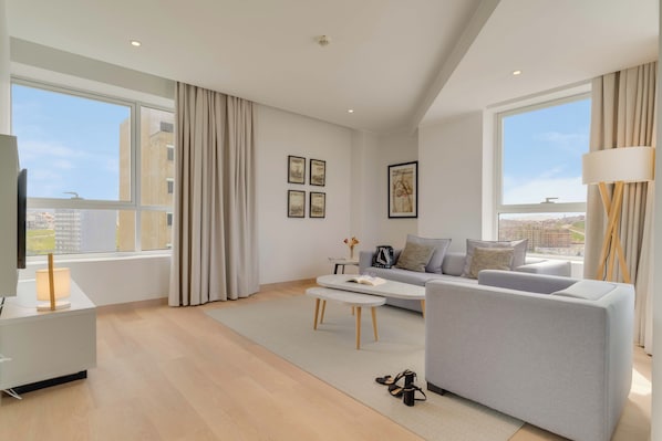 A modern living room with a grey sofa, a coffee table, and a TV on a stand. There are large windows with curtains, a city view, and a lamp on a side table.