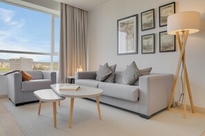 Uma sala de estar moderna com um sofá cinza, uma mesa de centro e um abajur. Há quadros na parede e vista da cidade pela janela.