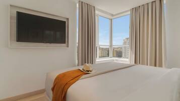 Habitación de hotel con una cama grande, un televisor montado y vistas a la ciudad a través de amplios ventanales.