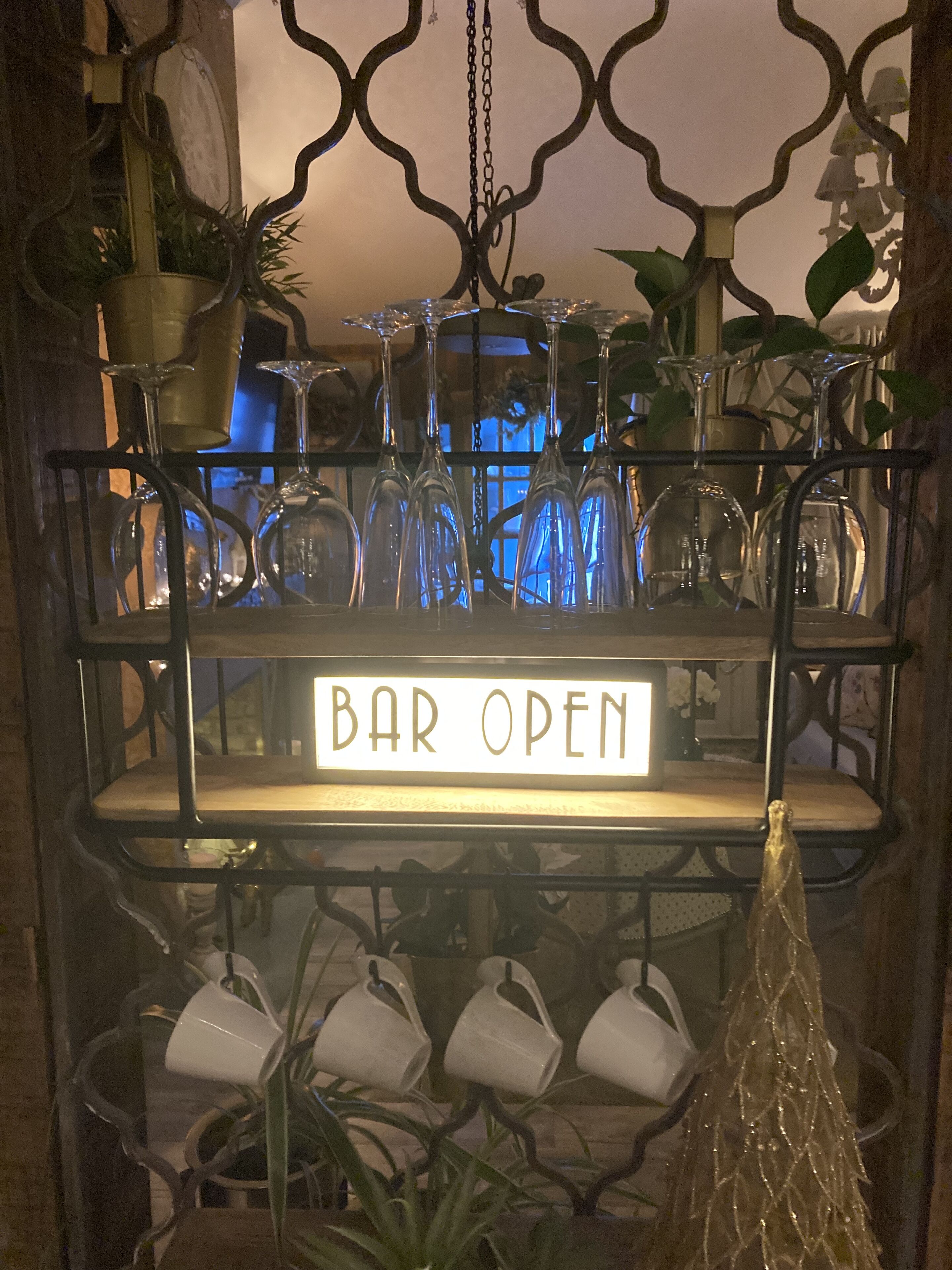 A bar area with a 'BAR OPEN' sign, shelves with wine glasses, and potted plants.