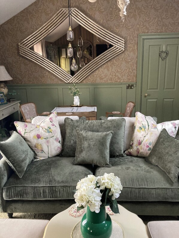 Un salon chaleureux avec un canapé vert, des coussins à motifs floraux, un vase vert rempli de fleurs et un miroir géométrique original accroché au mur.