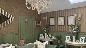 A cozy living room with a green sofa, a round coffee table, and a chandelier. There is a door with a heart-shaped handle, a small table with framed pictures, and a potted plant.