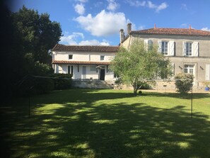 Una casa de dos pisos con techo de tejas, una puerta principal y un pequeño jardín con un árbol y césped.