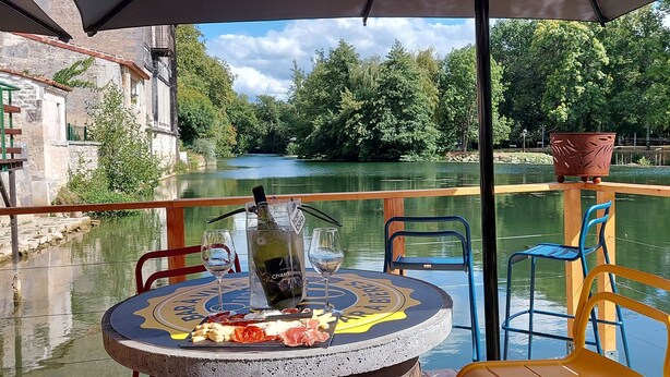 Ein Tisch mit einer Weinflasche, einem Glas und einem Teller Essen auf einem Bootsdeck mit Blick auf einen Fluss und grüne Landschaft.