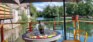 Una mesa con una botella de vino, una copa y un plato de comida en la cubierta de un barco, con vistas a un río y vegetación.