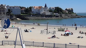 Ein Strand mit sonnenbadenden und schwimmenden Personen, ein Schild mit einer Karte und Gebäude im Hintergrund.