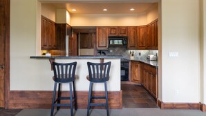 A kitchen with wooden cabinets, a central island with two bar stools, a microwave, and a sink. - Grand View 3330: Top Floor Condo w/ Spectacular Teton Views- Air Conditioned! (Jackson)