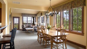 A spacious dining area with a large wooden table and chairs, a chandelier, and large windows offering a view of greenery. - Grand View 3330: Top Floor Condo w/ Spectacular Teton Views- Air Conditioned! (Jackson)