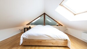 A bedroom with a sloped ceiling, a bed with white bedding, a small bedside table, and a wall-mounted light fixture. - Spacious Duplex Close to all Cultural Sites (Brussel)