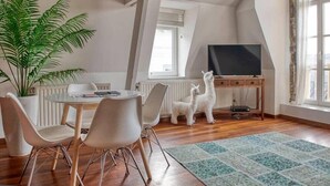 Ein Zimmer mit einem Esstisch, Stühlen, einer Topfpflanze, einem Fernseher auf einem Schrank und einem Fenster.