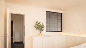 Une chambre à coucher avec un lit, une armoire blanche, une plante en pot et une fenêtre avec des stores.