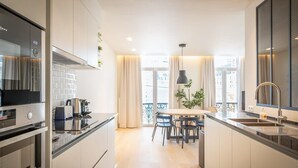 Une cuisine moderne avec des armoires blanches, une cuisinière en acier inoxydable, un coin repas avec une table et des chaises, et une vue sur la ville grâce à de grandes fenêtres.
