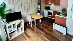 Ein kompakter Wohnraum mit Holzboden, einem Esstisch, einem Fernseher auf einem Schrank, einer Küchenzeile mit Schränken und einem Heizkörper.