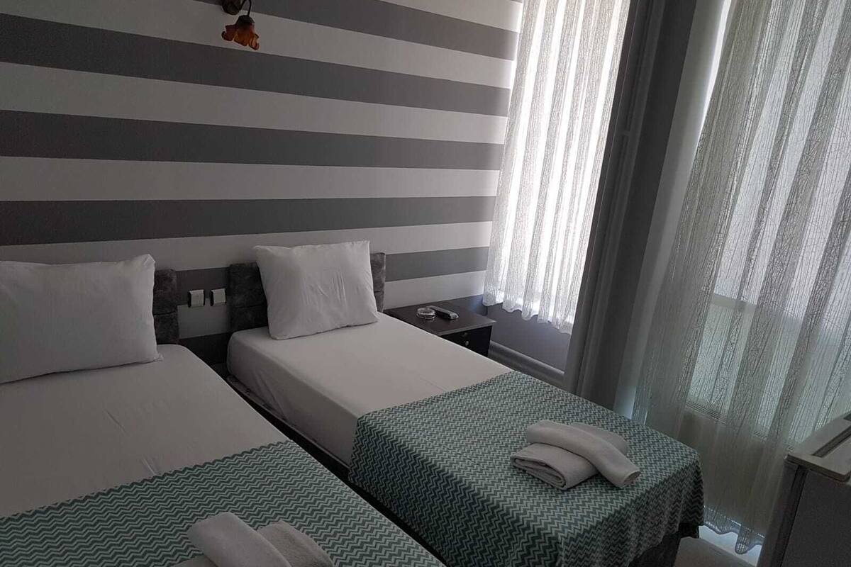 Habitación de hotel con dos camas, cabecera a rayas, una mesita de noche y una ventana con cortinas traslúcidas.