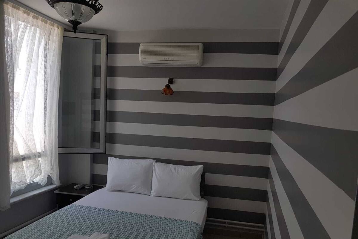 Una habitación de hotel con cama, mesita de noche, espejo, unidad de aire acondicionado y ventana con cortinas translúcidas.