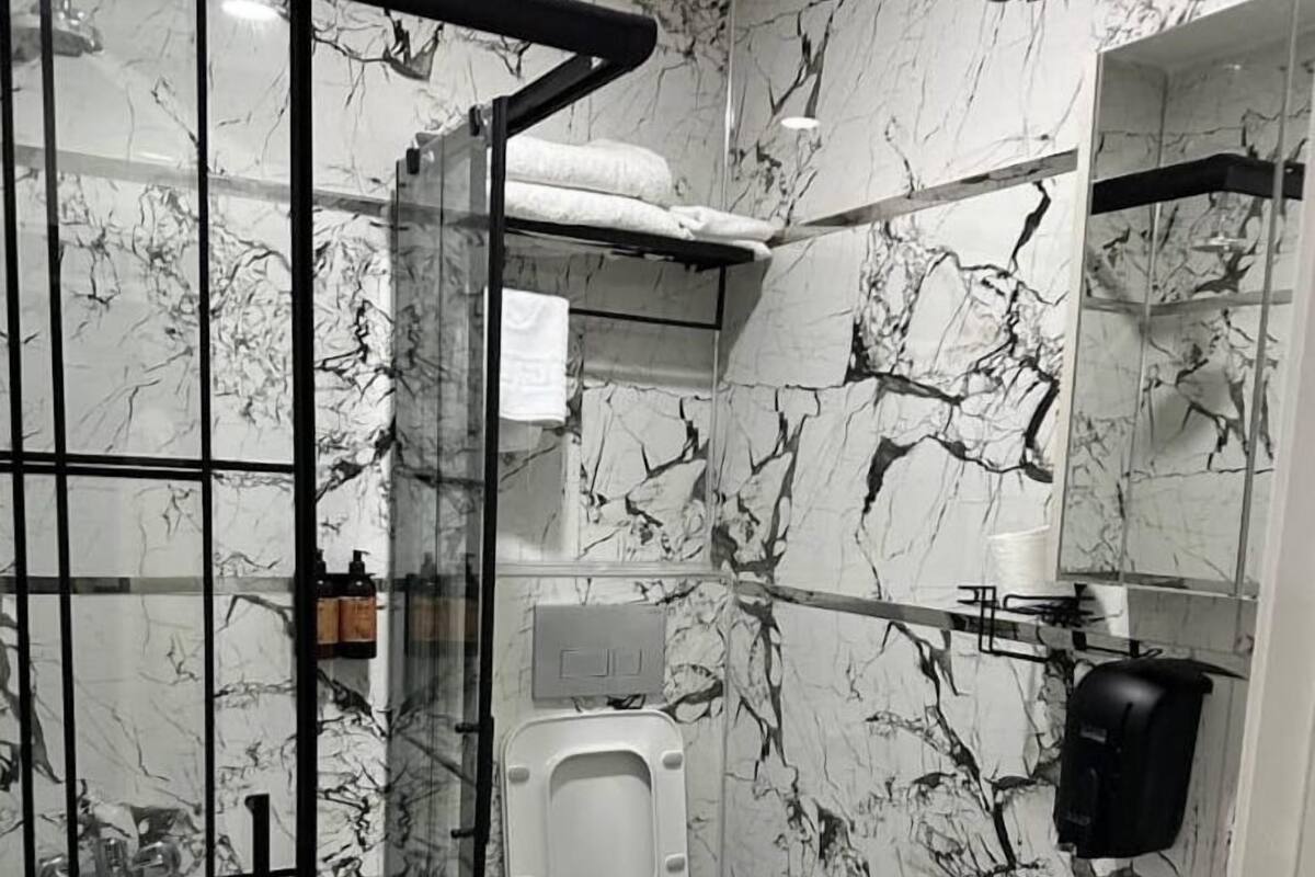 Un baño con pared de diseño de mármol, cabina de ducha de vidrio, inodoro, lavamanos y espejo.