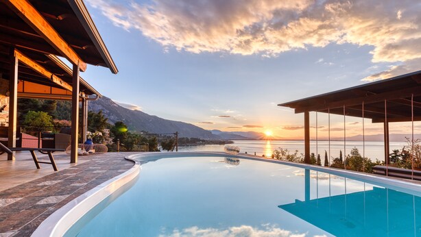 Une piscine avec vue sur le coucher de soleil sur l’eau, un patio couvert et un paysage montagneux en arrière-plan.