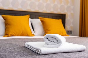 Ein ordentlich bezogenes Bett mit weißen und gelben Kissen, ein gefaltetes weißes Handtuch und eine graue, strukturierte Bettdecke.