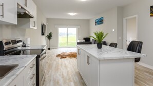 Una cocina moderna con armarios blancos, encimera de mármol y una isla grande. Hay una zona de comedor con una mesa y sillas, y vistas a un paisaje verde a través de una puerta corrediza de vidrio.