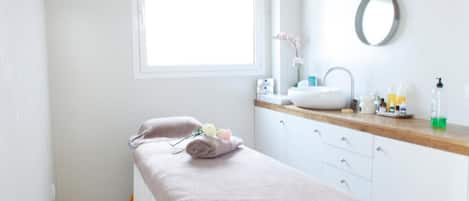 A clean, well-lit room with a bed, a bedside table with toiletries, a window, and a mirror on the wall.