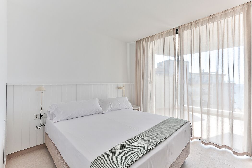 Una habitación de hotel con una cama grande, una mesita de noche, una lámpara y cortinas traslúcidas.