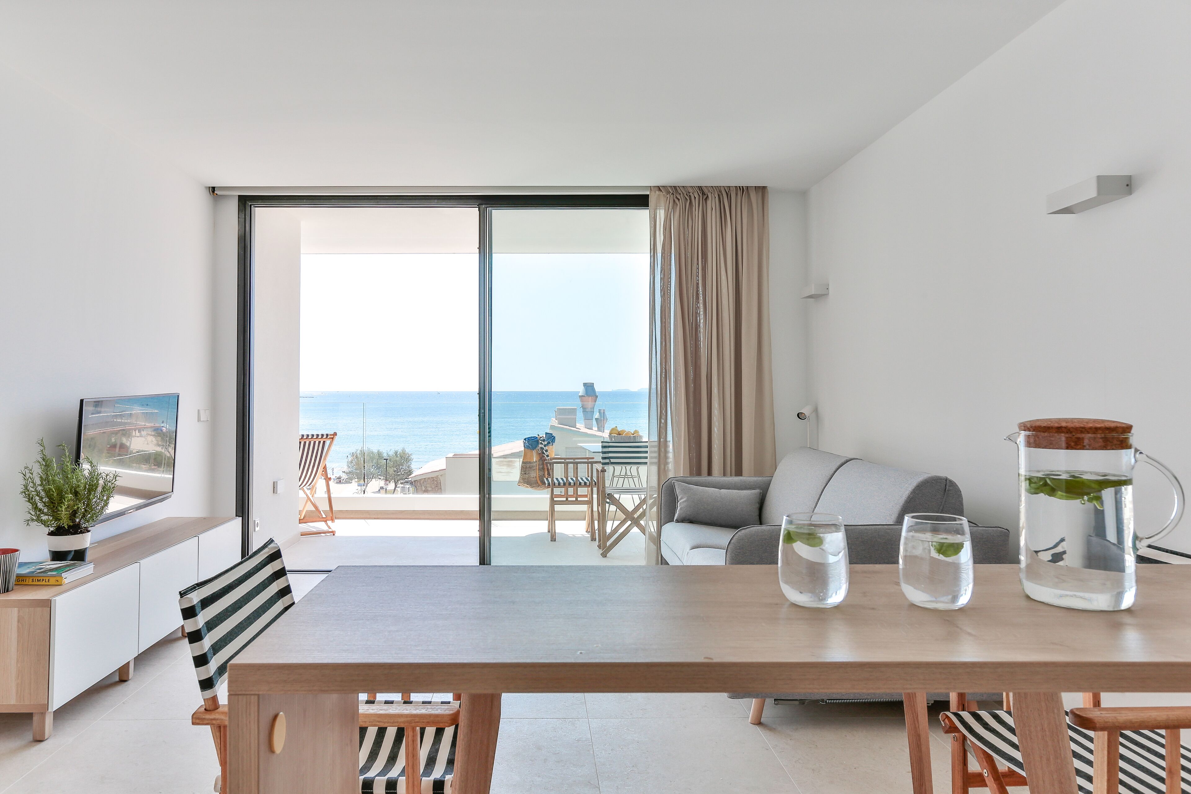 Un salon moderne doté d’une grande porte-fenêtre coulissante donnant sur un balcon avec vue sur la mer. On y trouve une table à manger avec des chaises, un canapé et une table basse avec une carafe et des verres.