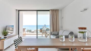 Una sala de estar moderna con un amplio ventanal corredizo que da a un balcón con vistas al mar. Hay una mesa de comedor con sillas, un sofá y una mesa de centro con una jarra y vasos.