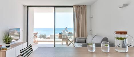 Un salon moderne doté d’une grande porte-fenêtre coulissante donnant sur un balcon avec vue sur la mer. On y trouve une table à manger avec des chaises, un canapé et une table basse avec une carafe et des verres.