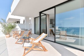 A modern outdoor patio with wooden lounge chairs and a table, a glass door leading to a kitchen, and a view of the sea. - ECO-APARTMENTS LA MARINEDA & HOTEL THALASSA SPORT & SPA (Roses)