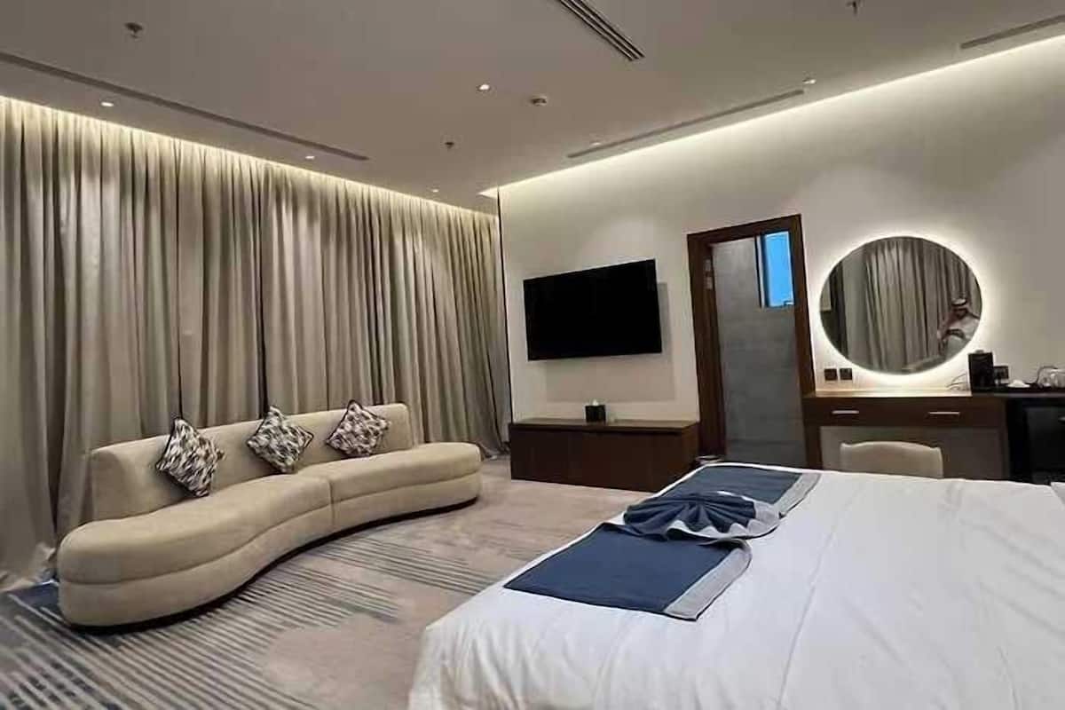 Una habitación de hotel moderna con un sofá curvo, una cama grande, un televisor de pantalla plana, un espejo redondo y un escritorio con lámpara.