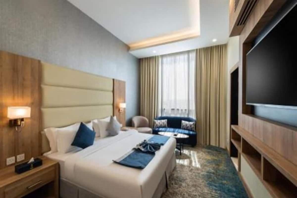 Quarto de hotel com uma cama grande, uma escrivaninha com abajur, um sofá e uma televisão.