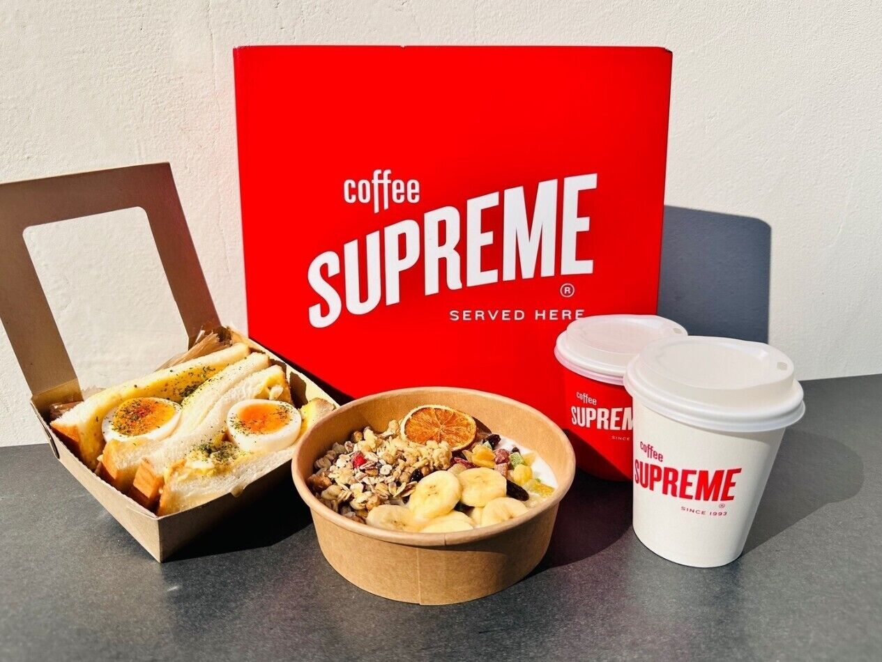 Un ensemble pour le petit-déjeuner comprenant un sandwich, un bol de céréales aux fruits et deux tasses à café, le tout portant la marque « Coffee Supreme ».