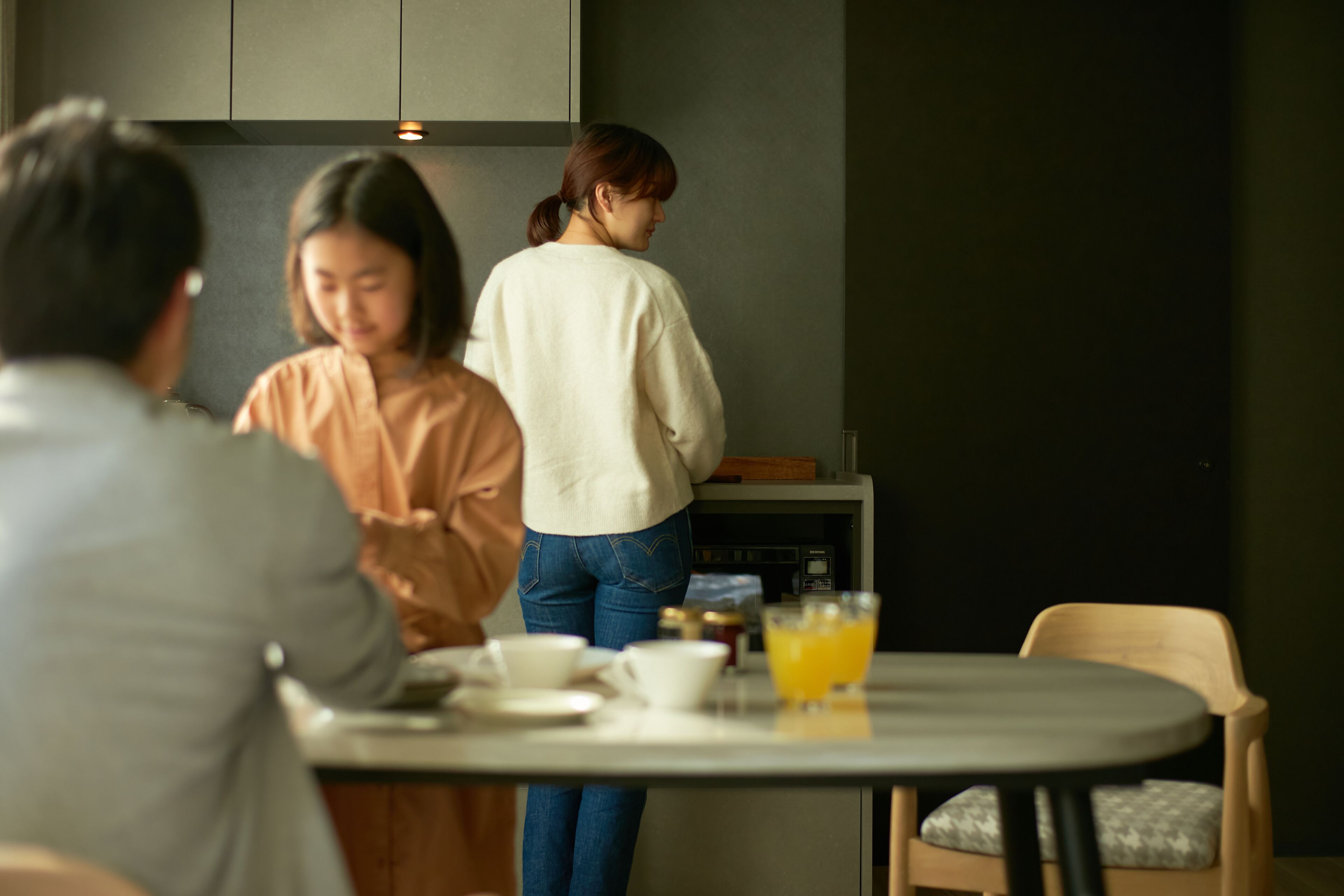 Un coin repas avec une table dressée pour deux personnes, une personne debout près du comptoir et une chaise avec un coussin.