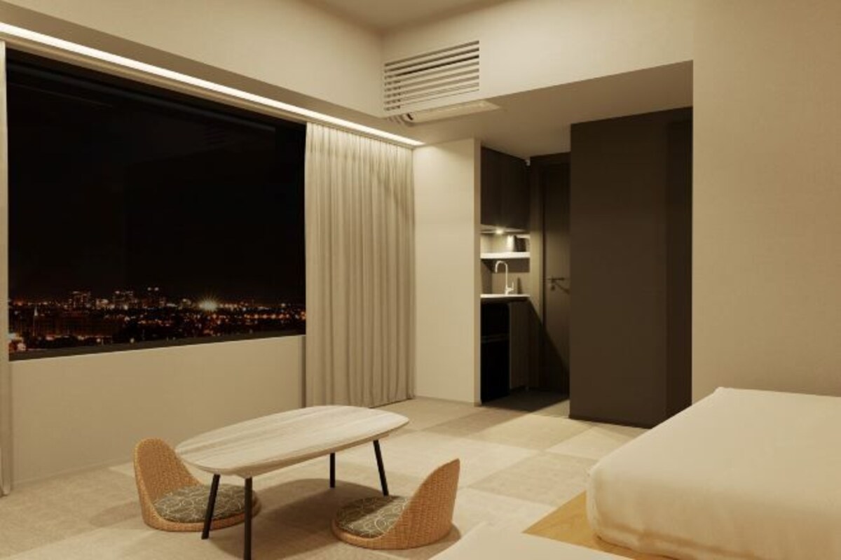 A modern hotel room with a large window offering a city view, a small dining area, and a separate sleeping area.