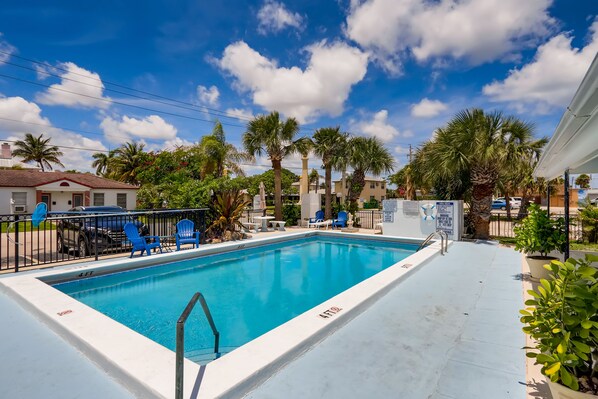 Une piscine rectangulaire dont la profondeur est indiquée par un repère à 4 pieds, entourée d’une terrasse blanche, de chaises bleues et de palmiers.