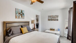 Quarto com cabeceira de madeira, cama com roupa de cama branca e cinza, ventilador de teto e duas pinturas penduradas na parede.