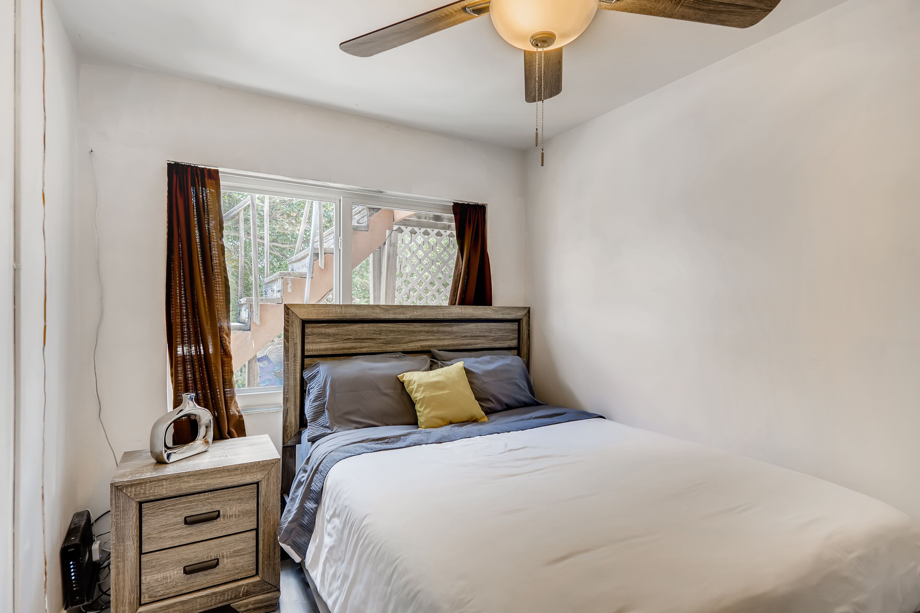 A bedroom with a wooden headboard, a bed with grey and yellow pillows, a bedside table with a radio, and a window with curtains.