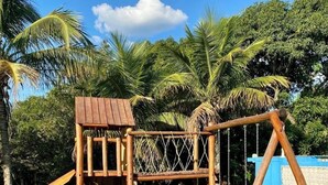 Une structure de jeux en bois, avec un toboggan et des balançoires, entourée de pelouse et de palmiers.
