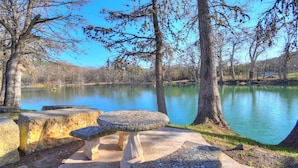 Un cadre paisible au bord d’un lac, avec un banc et une table en pierre, entourés d’arbres et sous un ciel dégagé.