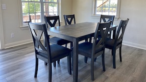 Un espace repas comprenant une table rectangulaire et six chaises, de grandes fenêtres à cadres blancs et un plancher en bois.