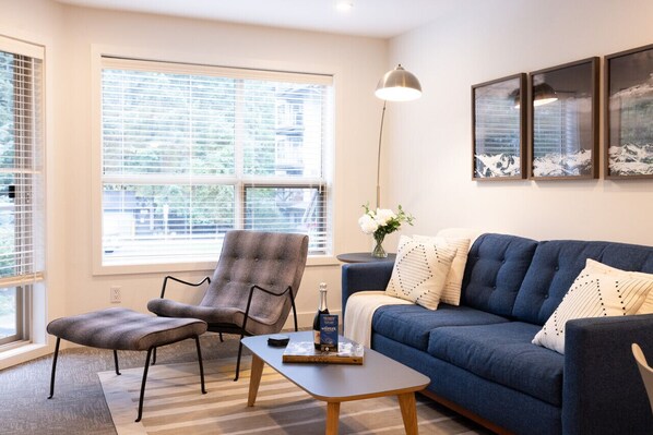 A living room with a blue sofa, a grey armchair, and a wooden coffee table. There is a large window with blinds, a floor lamp, and framed artwork on the wall.