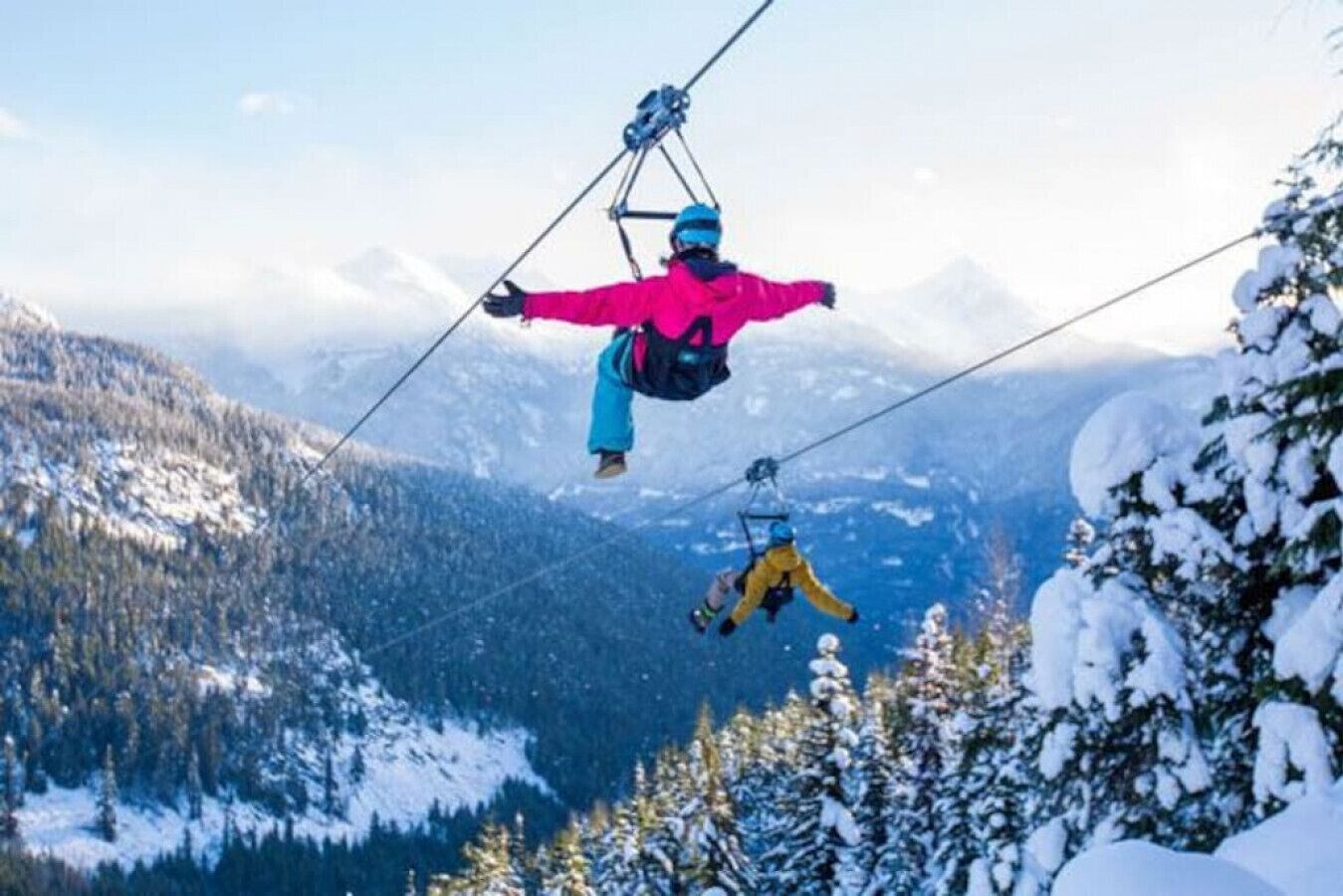 Una persona con chaqueta rosa y pantalones azules suspendida en un cable, con un paisaje montañoso nevado al fondo.