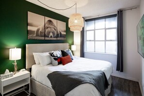 A hotel room with a large bed, a headboard, a bedside table with a lamp, a window with curtains, and a wall art piece.
