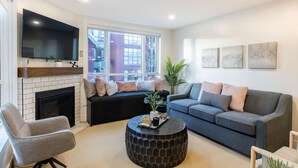 A modern living room with a fireplace, a large flat-screen TV, a grey sofa, a round coffee table, and a window with a view of buildings.